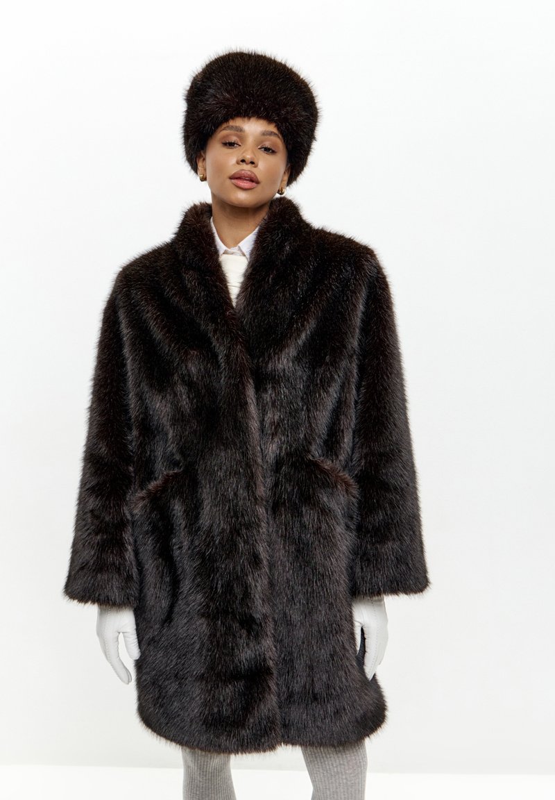 Brun faux fur frakke med en bred krave, to sidelommer og matchende pels hat. Teksturen er tyk og fyldig, hvilket skaber et varmt udseende.