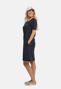 Rochie scurtă de marină cu mâneci scurte, buzunare laterale, decolteu rotund și un logo mic. Asortată cu o pălărie albă și papuci tip flip-flop. Material neted și casual.