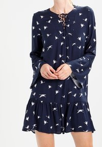 Navy sininen mekko, jossa on kellosuut, nauhakäyttöinen kaula-aukko ja lintukuosi. Leveä helma, jossa on kerroksellinen röyhelöyksityiskohta. Silkkinen kangas.