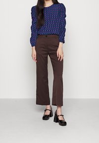 Top a pallini viola e verde con maniche a sbuffo abbinato a pantaloni marroni su misura e scarpe con piattaforma nere. Texture lisce, design aderente.