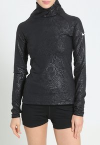 Mujer que lleva una camiseta negra de manga larga y cuello alto con un discreto estampado de serpiente y pantalones cortos negros, de pie con las manos relajadas a los lados.