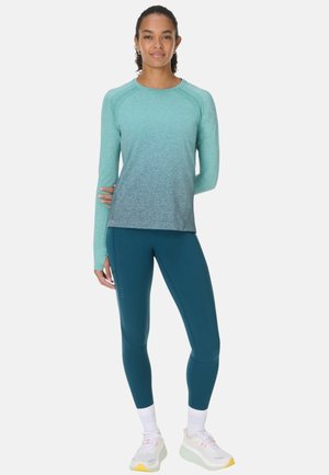Vrouw staand, draagend een teal gradient lange mouwen shirt, donkere teal legging, witte sokken en witte sneakers met gele zolen.