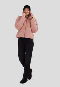 Różowa puchowa kurtka z sztruksu z zamkiem z przodu, z wysokim kołnierzem. W zestawie z czarnymi spodniami o prostym kroju i czarnymi sneakersami.