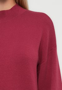 Maglione magenta con collo alto, realizzato in un morbido tessuto testurizzato. Presenta spalle scese e maniche larghe per un fit rilassato.