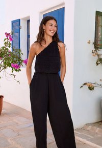 Sort jumpsuit med en struktureret et-skulder-design, vide ben og sidelommer, med lagdelt hæklet detalje ved kroppen.