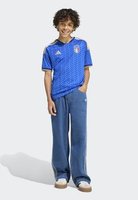 Blå fotbollströja med texturerat mönster och gulddetaljer, kombinerad med löst sittande blå denimbyxor och vita sneakers. Justerbar krage för passform.