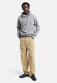 Felpa grigia con cappuccio e logo in rilievo, abbinata a pantaloni beige a gamba dritta e mocassini neri. Tessuto morbido, vestibilità rilassata.
