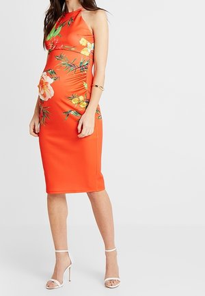 Zwangere vrouw draagt een mouwloze, aansluitende oranje midi-jurk met bloemenprint, gecombineerd met witte sandalen met hoge hak en een gouden armband.