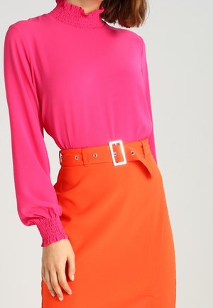 Femme portant un chemisier à manches longues rose vif avec des poignets et un col froncés, associé à une jupe orange taille haute avec une large ceinture.