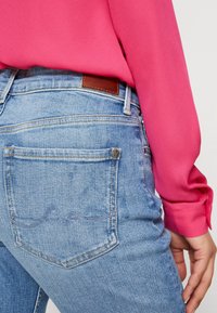 Jeans de mezclilla azul claro con una etiqueta de cuero en la parte posterior, que cuentan con dos bolsillos traseros y detalles de costura sutiles. Combinados con una blusa rosa.