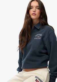 Sudadera recortada de color azul marino con cuello tipo camisa, que presenta el texto "Superdry Athletics" y un emblema. Tejido suave y texturizado.