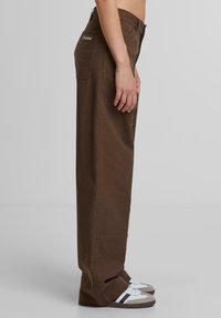 Braune weite Hosen aus Baumwolltwill mit Seitentaschen und Logo. Kombiniert mit grauen und weißen Sneakers.