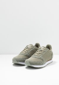 Grüne Sneakers aus Mesh- und Wildledermaterialien, mit strukturiertem Design, weißen Sohlen und braunem Akzent, ausgestattet mit klassischen Schnürsenkeln und abgerundeter Zehenpartie.