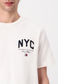 White cotton t-shirt featuring navy embroidered text "NYC" and "ALL AMERICAN CLASSIC GOODS" with Tommy Hilfiger logo detail. Short sleeves.