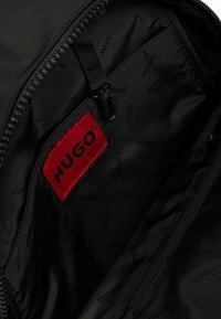 Black textile interior of a bag featuring a prominent red label with "HUGO" printed in black, alongside repeating black "HUGO" patterns.