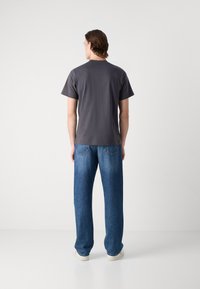 Grå bomull t-skjorte med rund hals, kombinert med løstsittende blå denimjeans og hvite joggesko, sett fra baksiden.