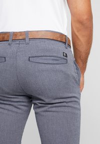 Pánské tmavě modré herringbone kalhoty s hnědým koženým opaskem. Obsahují dvě zadní kapsy a mají štíhlý střih s jemnou texturou.