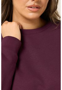 Burgundy sweatshirt med rund halsringning, ribbad ärmavslut och mjuk tygtextur, delvis visad på en modells överkropp.