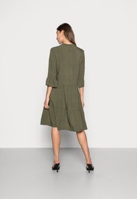 Vestido verde-oliva com mangas até ao cotovelo, saia em camadas e padrão de pequenas bolinhas. Apresenta um decote redondo e punhos de manga com babados.