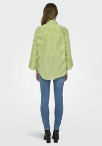 Camisa verde de rayas oversize con un dobladillo asimétrico y mangas anchas, combinada con jeans ajustados azules y botas de tobillo de tacón ancho negras.