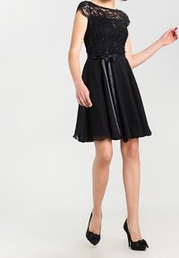 Svart klänning med spetsliv, korta ärmar och ett band i midjan; svag kjol som slutar ovanför knäet. Svarta klackar med rosett detaljer.