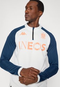 Αθλητική μπλούζα μακρυμάνικη σε λευκό και ναυτικό μπλε. Διαθέτει λαιμόκοψη με φερμουάρ, πορτοκαλί λογότυπο "INEOS", έμβλημα Kappa σε λευκό και εφαρμοστά μανίκια. Μαλακή υφή υφάσματος.
