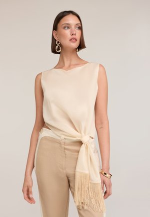 Femme portant un haut beige sans manches noué avec un détail à franges, associé à un pantalon beige taille haute et de grandes boucles d'oreilles dorées.