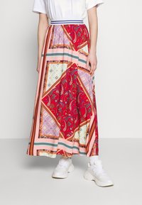 Falda larga y estampada en rojo, rosa y crema. Presenta diseños de paisley y geométricos, con una cinturilla y rayas en contraste. Combinada con zapatillas blancas.
