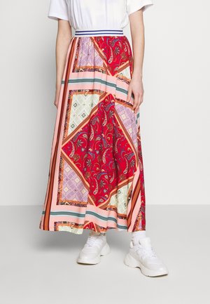 Jupe longue à motifs en rouge, rose et crème. Présente des motifs paisley et géométriques, avec une taille contrastante et des rayures. Portée avec des baskets blanches.