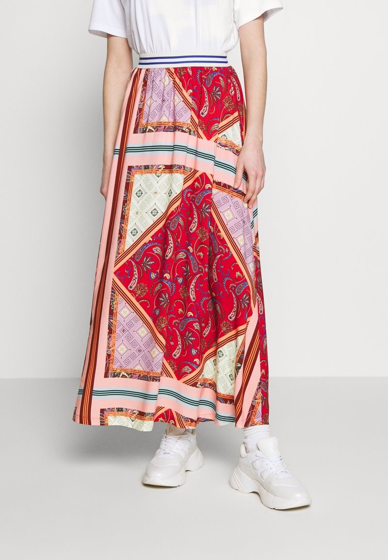 Falda larga y estampada en rojo, rosa y crema. Presenta diseños de paisley y geométricos, con una cinturilla y rayas en contraste. Combinada con zapatillas blancas.