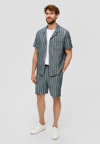 Herren Kurzarmhemd mit blauen und weißen Streifen, kombiniert mit passenden Shorts. Mit Knopfverschluss und lässigem Schnitt. Getragen mit weißen Sneakers.