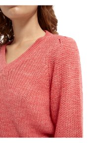 Suéter de punto color coral con cuello en V, puños acanalados y hombros ligeramente abullonados. Textura suave con un ajuste holgado, que revela un tejido abierto.