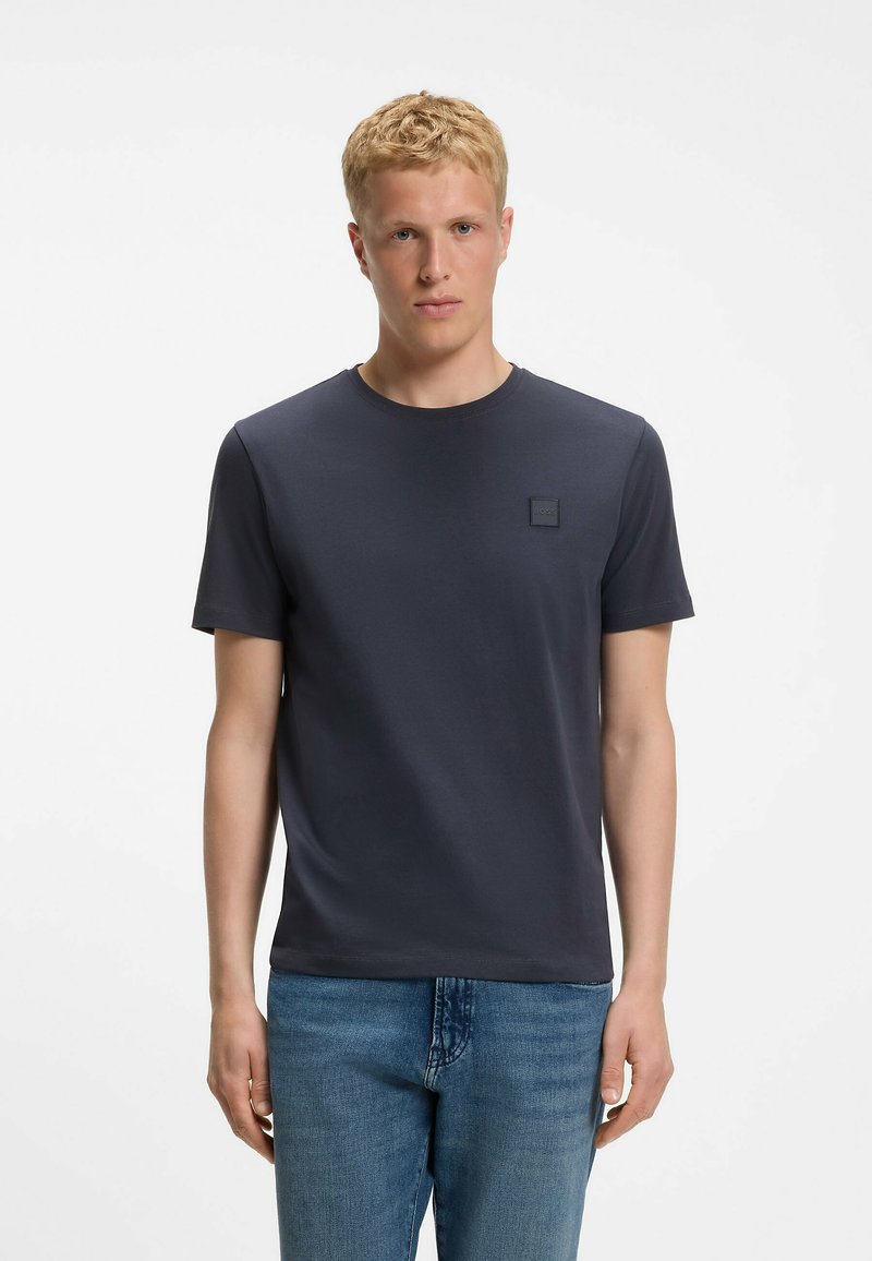 Homme blond portant un t-shirt bleu marine uni à manches courtes et un jean bleu, debout devant un fond blanc.