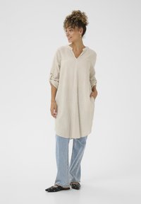 Tunica a righe a maniche lunghe in beige e crema, con scollatura a V e tasche laterali, abbinata a jeans blu chiaro e scarpe nere slip-on.