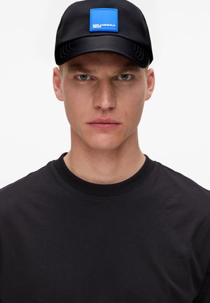Gorra negra con un parche de logo cuadrado azul, combinada con una camiseta negra de cuello redondo. La gorra presenta un visera curva y acentos de costura.