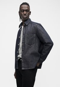 Tmavě modrá denimová košile s zapínáním na knoflíky, špičatým límečkem a náprsní kapsou. Natažená přes světle šedý svetr a černé kalhoty.