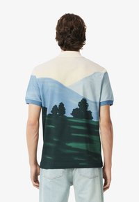 Lyhytohiainen polo-paita, jossa on maisemakuva sinisistä vuorista ja tummanvihreistä puista. Paita on valmistettu pehmeästä kankaasta, joka on sileä kosketukseltaan.