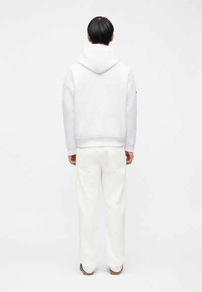 Άτομο με κοντά μαύρα μαλλιά που φοράει λευκό φούτερ με κουκούλα και λευκό παντελόνι, όρθιο σε λευκό απλό φόντο.