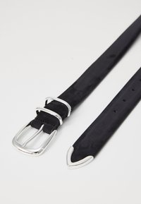 Cinturón de ante negro con hebilla y herrajes de metal plateado. Presenta múltiples agujeros para ajuste y un diseño de extremo puntiagudo.