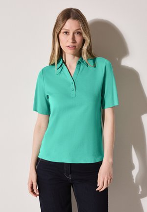 Short-sleeve polo shirt in vibrant turquoise, made from soft fabric. Features a pointed collar and three-button placket. Paired with dark jeans.