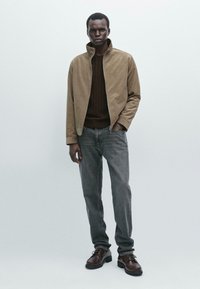Giacca in corduroy color tan con colletto alto, maglione marrone a maglia, jeans grigi e stivali marroni con lacci. Il modello è in piedi contro uno sfondo neutro.