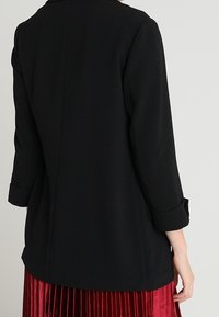 Žena v černém saku s vyhrnutými rukávy a červené skládací sukni, viděná zezadu na jednobarevném pozadí.