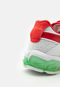 Vit sportskor med röd hälflik som har Coca-Cola-logotyp, röd rand och detalj med genomskinlig grön dämpad sula.