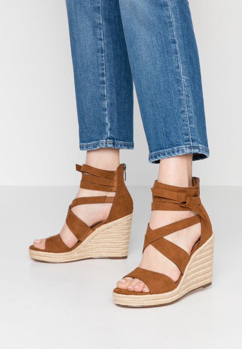 Pieds portant des sandales compensées en daim marron avec des brides à la cheville, associées à un jean denim bleu retroussé sur un fond clair.