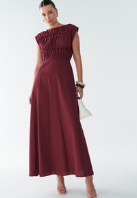 Vínové maxi šaty s krátkymi rukávmi, nazberaným pásom, plynúci textilom a hladkou textúrou. Nosené s ľahkou kabelkou a sandálmi na remienky.