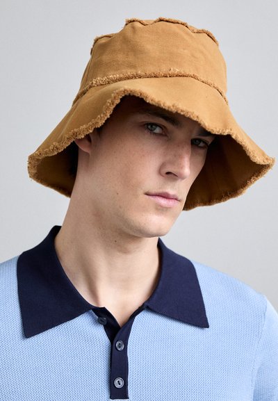Brązowy kapelusz typu bucket z postrzępionymi brzegami, wykonany z materiału. Noszony przekrzywiony, w połączeniu z jasnoniebieską, teksturowaną koszulą polo z granatowym kołnierzykiem.