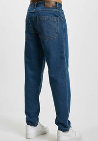 Vaqueros de mezclilla azul con un corte relajado, que cuentan con una etiqueta de cuero en la cintura, dos bolsillos traseros y tobillos enrollados, combinados con zapatillas blancas.