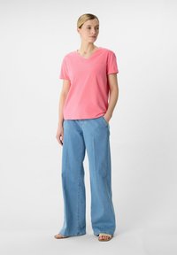 Różowa bawełniana koszulka z dekoltem w serek i krótkim rękawem, zestawiona z jasnoniebieskimi spodniami typu wide-leg. Modelka stoi w luźnej pozie, z jedną ręką w kieszeni.