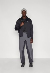 Casaco acolchoado preto sobre uma camisa preta, calças cinza com costuras distintas e sapatos pretos. Acessório: gorro cinza claro.