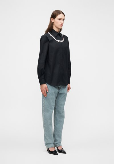 Γυναίκα όρθια, φορεί ένα μαύρο πουκάμισο με μακριά μανίκια και λευκό σχέδιο κουμπιών κοντά στον γιακά, ανοιχτό μπλε τζιν και μαύρες μυτερές γόβες.